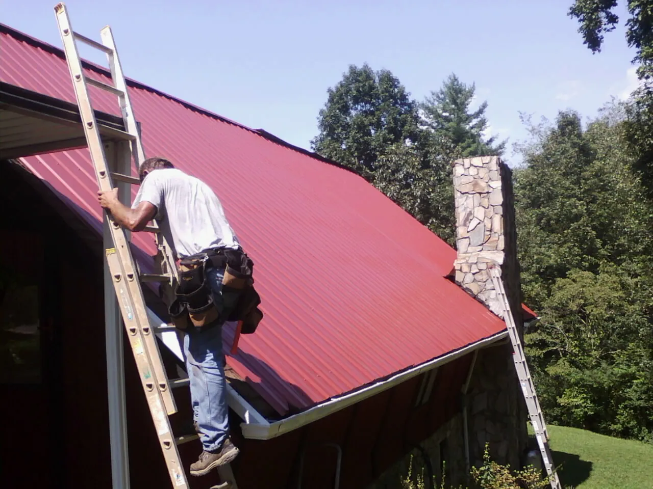 Roofer on a ladder beside a red metal roof and stone chimney — Soffit & Fascia Repair by Roofing Brothers in San Pablo